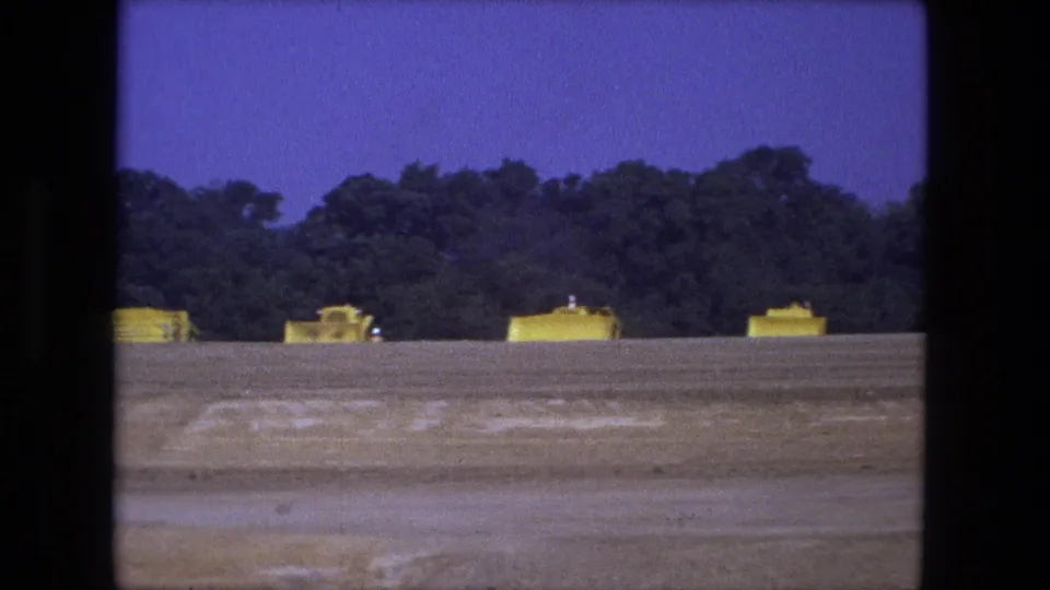 Wide 1974 Fort Wayne home-movie view of yellow construction vehicles lined up beyond a bare dirt field and tree line.