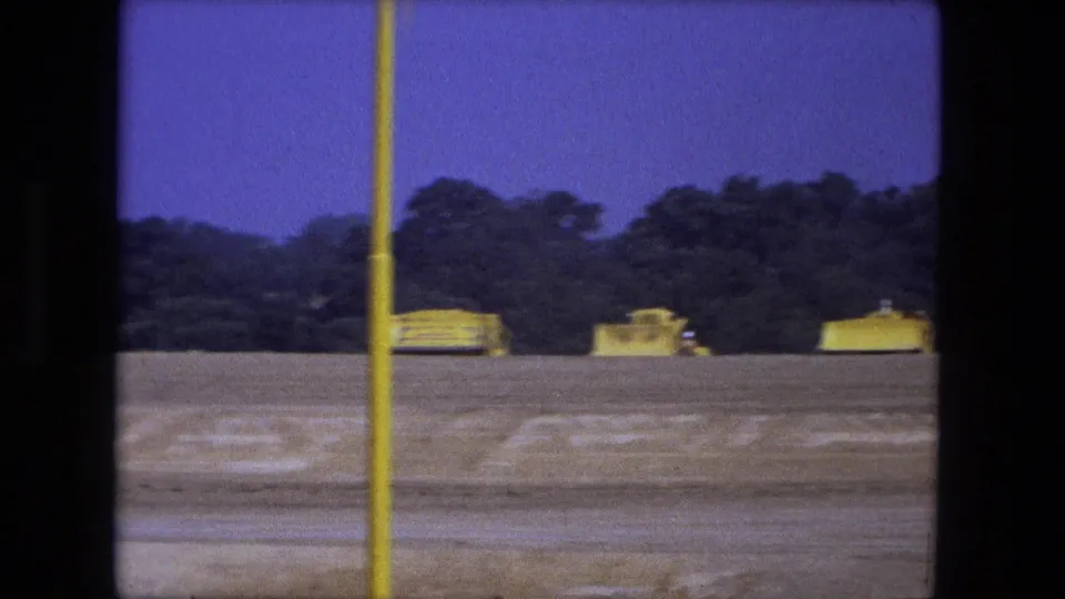 1974 Fort Wayne 8mm frame with distant yellow vehicles, a vertical yellow pole, trees, and a purple sky over a dirt field.