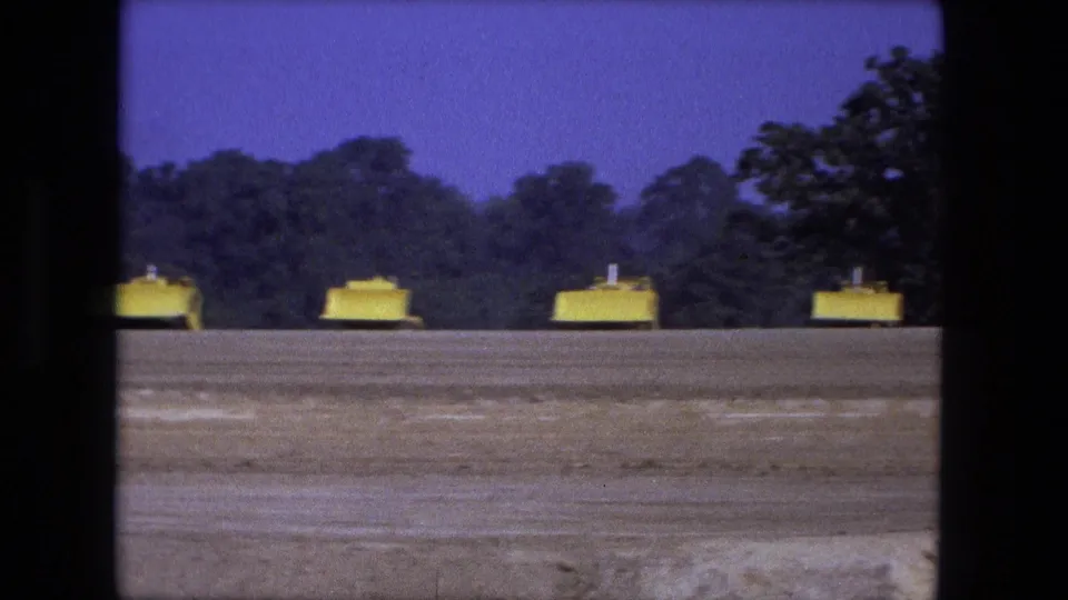 Wide 1974 Fort Wayne 8mm view of yellow vehicles along the far edge of a dirt field under a purple sky.