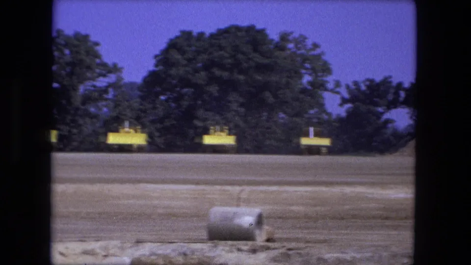1974 Fort Wayne frame with yellow construction equipment near trees and a concrete pipe lying in the foreground of the graded site.