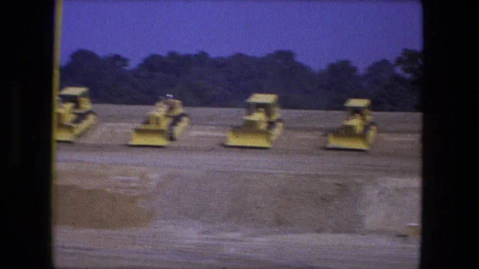1974 Fort Wayne frame showing yellow bulldozers aligned on a graded construction site, with dark trees and blue sky behind them.