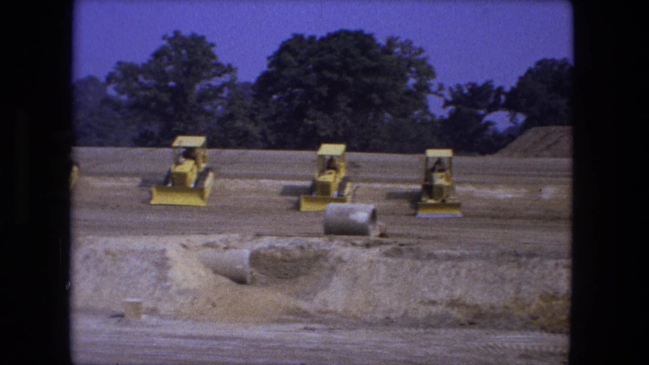 Yellow loaders move on a dirt site near concrete pipes and low piles of soil.