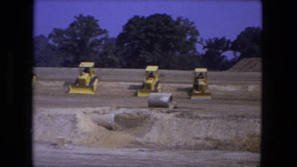 1974 Fort Wayne construction-site frame with yellow loaders, concrete pipes, soil piles, and trees beyond the graded field.