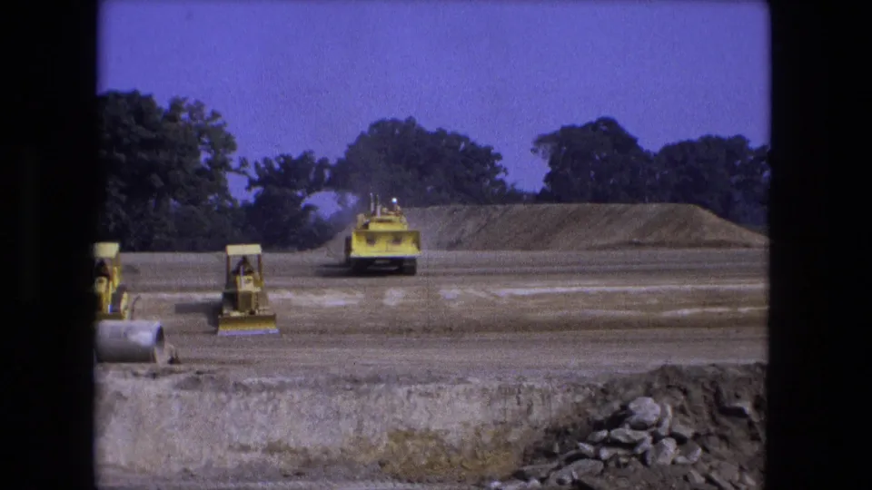 1974 Fort Wayne 8mm frame showing a loader facing the camera, another machine near concrete pipes, and a soil mound beyond.