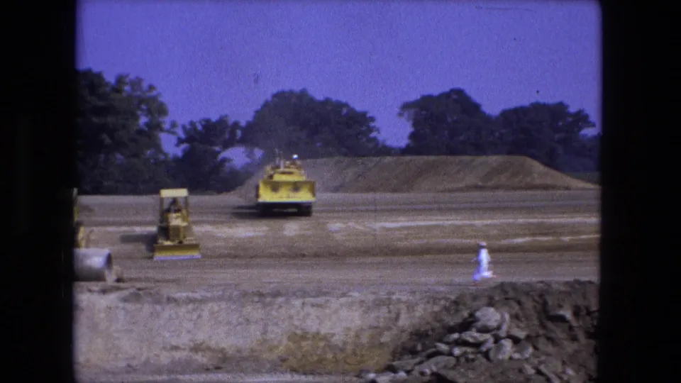 1974 Fort Wayne frame with a yellow loader on the graded field and a distant worker in light clothing crossing the site.