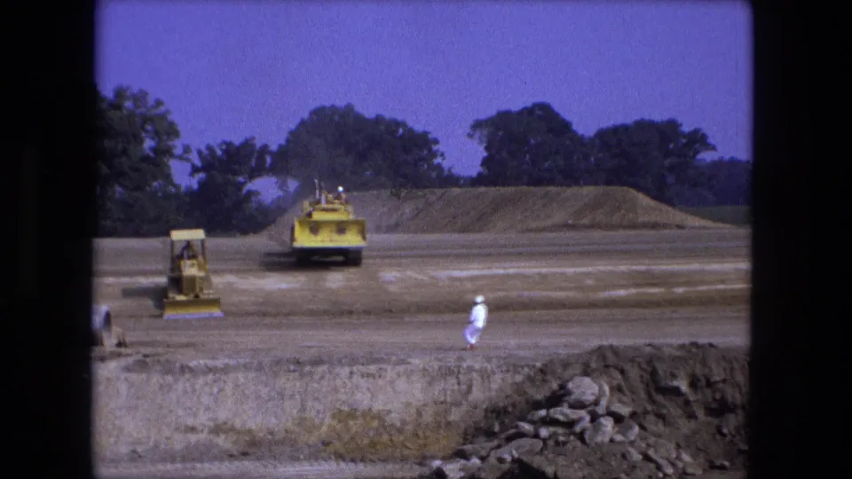 1974 Fort Wayne 8mm frame showing yellow earthmoving machines on a graded construction site with workers and trees in the background.