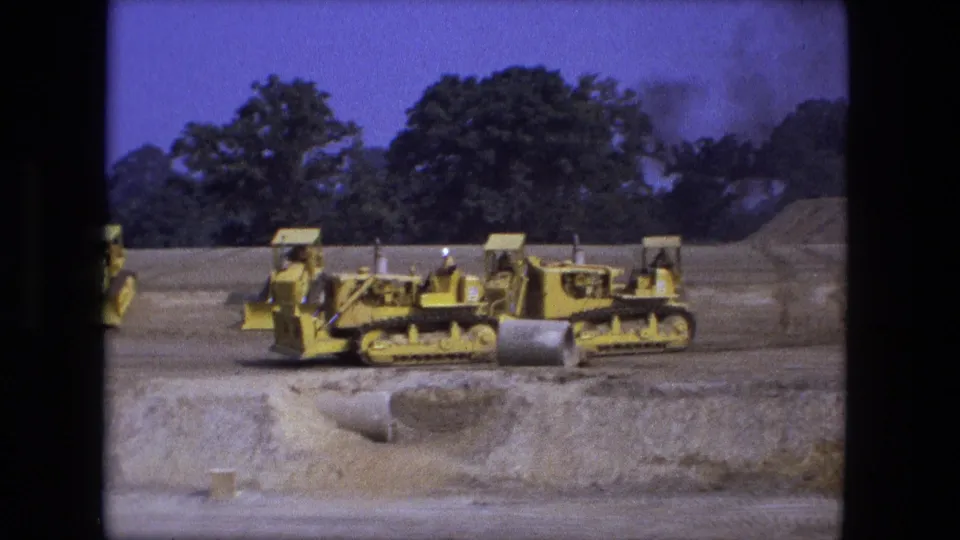1974 Fort Wayne 8mm frame of yellow tracked construction machines traveling across a dirt work zone with trees behind them.