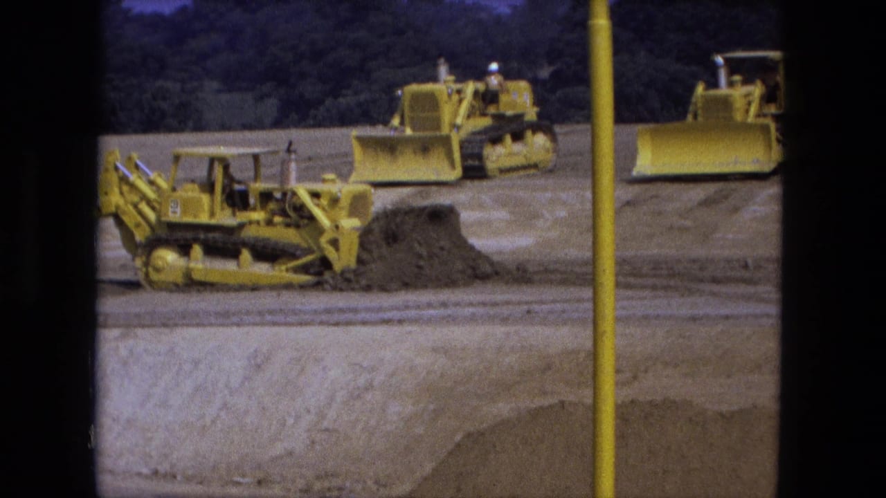 Yellow bulldozers work across a dirt excavation site with a pole and wooded background.