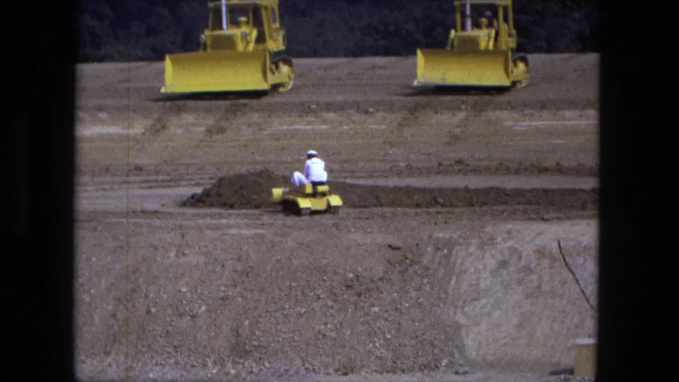 1974 Fort Wayne 8mm frame showing a distant worker operating a small yellow tracked machine on a dirt construction site.