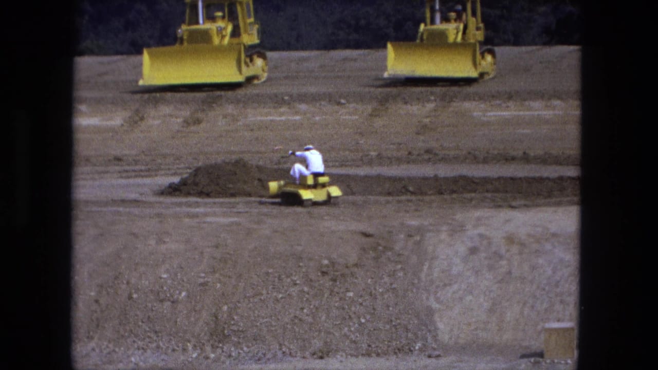 Worker in a white shirt rides a small yellow dozer on a sandy construction lot with two larger yellow dozers behind.
