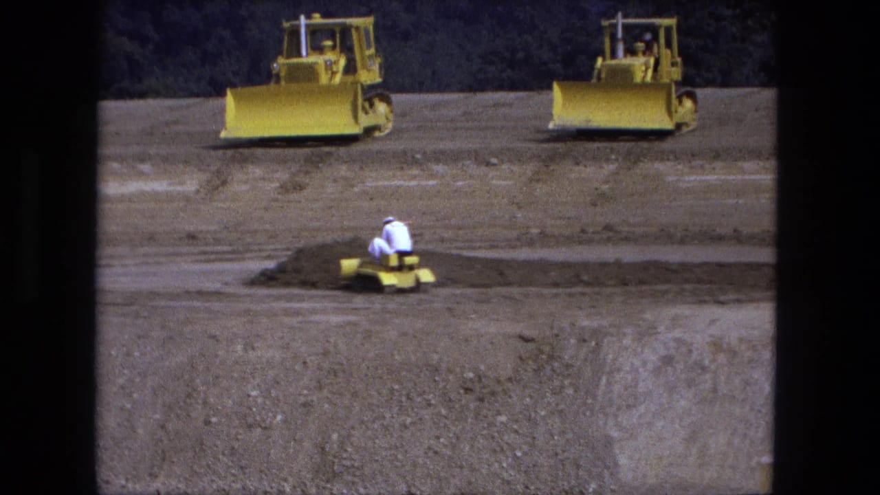 Fort Wayne construction-site frame from 1974 with a worker on a small yellow dozer and larger bulldozers on the open sandy lot.