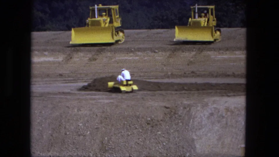 Fort Wayne construction-site frame from 1974 with a worker on a small yellow dozer and larger bulldozers on the open sandy lot.