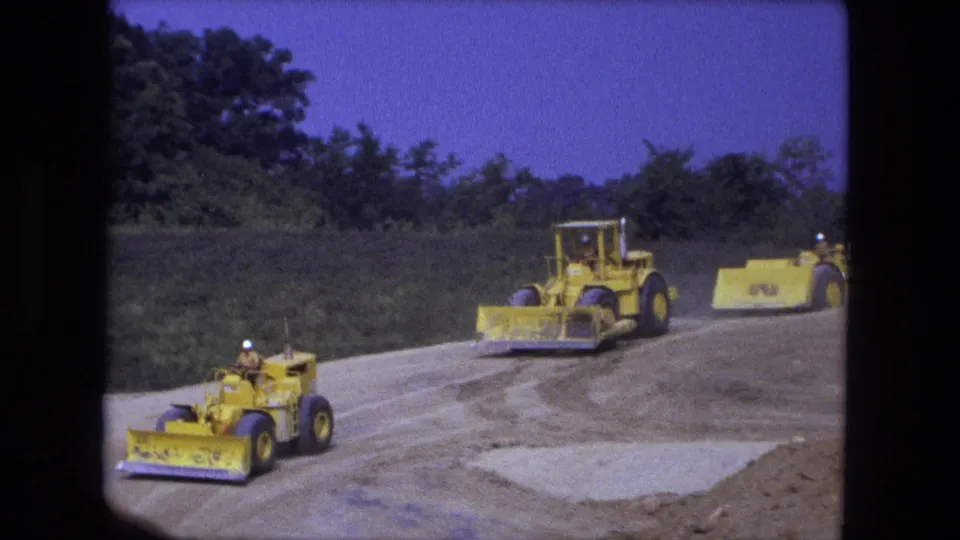 1974 Fort Wayne 8mm frame showing yellow construction machines rolling along a graded road beside trees under blue sky.