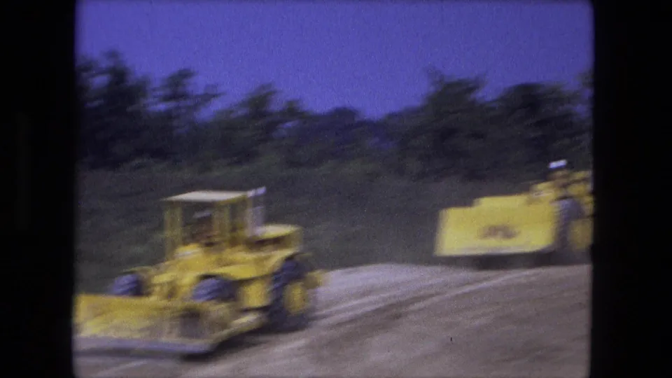 Motion-blurred 1974 Fort Wayne frame of yellow bulldozers moving along a graded construction road near a wooded edge.