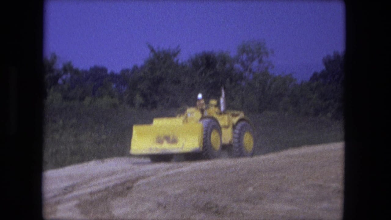Yellow earth mover driven by a hard-hat worker at a Fort Wayne construction site in 1974, seen from a distance on 8mm film.