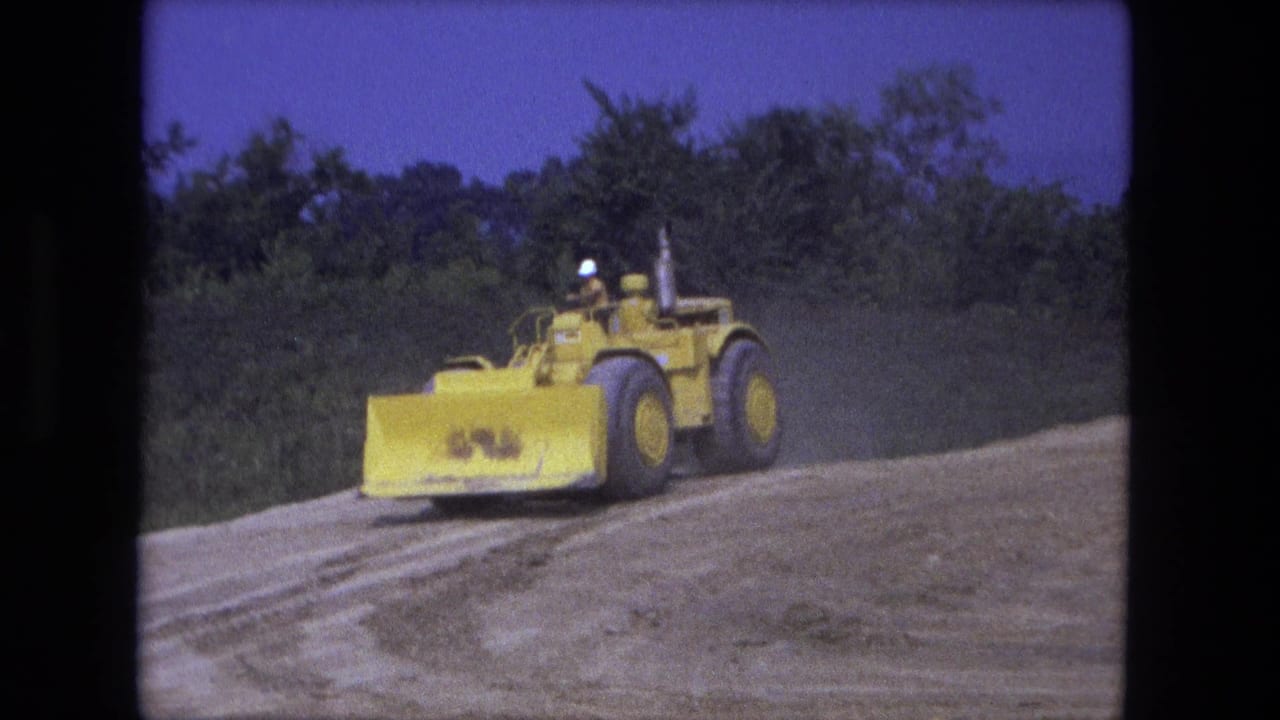 Yellow earth mover turns across a dirt surface near low trees.