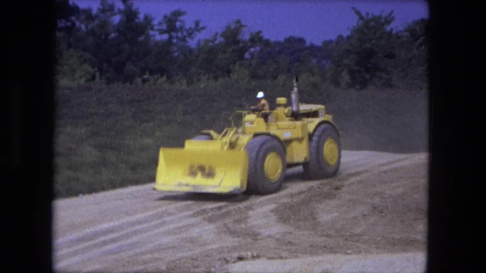 A yellow earth mover with a hard-hat operator drives past on the dirt construction site in Fort Wayne, Indiana.