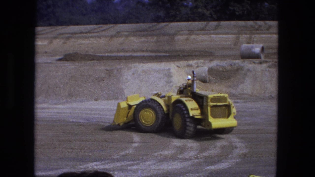 Yellow front-end loader on a graded dirt field with mounds and a tree line behind it.
