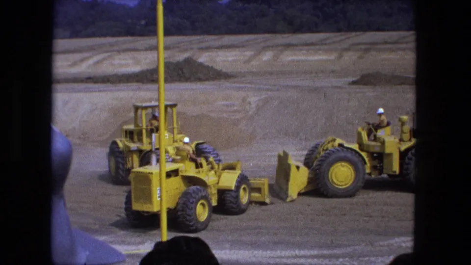1974 Fort Wayne 8mm frame showing a grader, a loader, and a yellow survey pole in the foreground.
