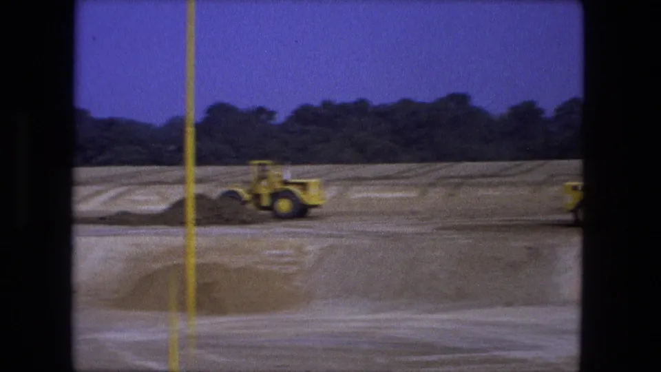 8mm Fort Wayne construction frame showing a blurred yellow loader moving past an earth pile and foreground pole.