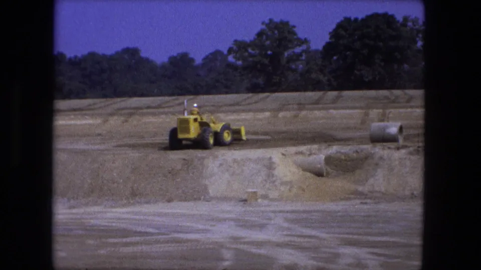 1974 Fort Wayne 8mm frame with a yellow loader grading near a dirt cut, visible pipe sections, trees, and blue sky.