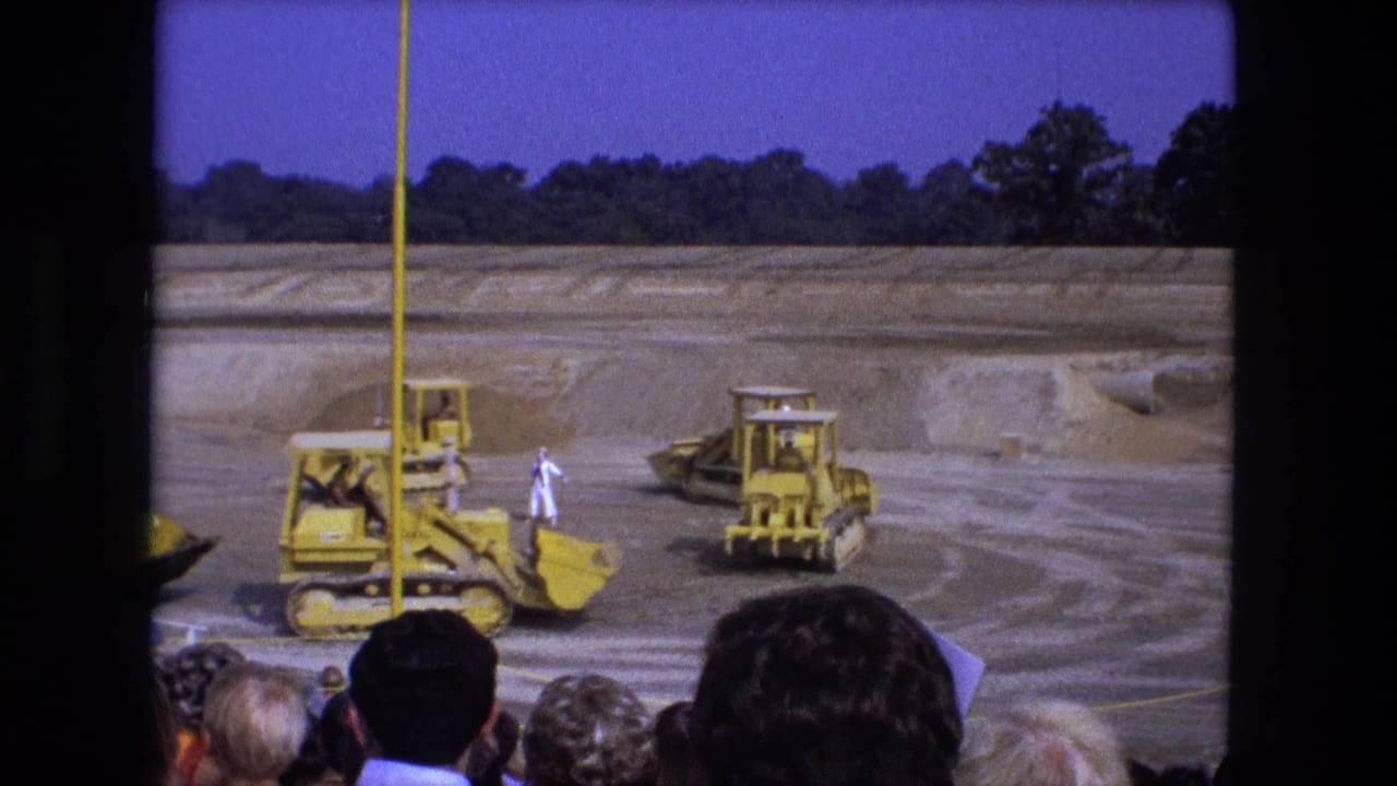 Yellow bulldozers and tractors move across a dirt construction site before spectators.