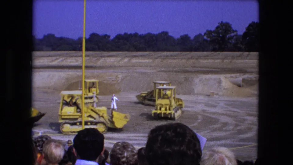 1974 Fort Wayne 8mm frame showing yellow construction machines crossing a graded dirt site as spectators watch from the foreground.
