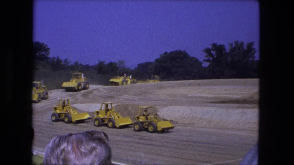 1974 Fort Wayne home-movie frame showing yellow front loaders and tractors passing spectators on a graded construction site.