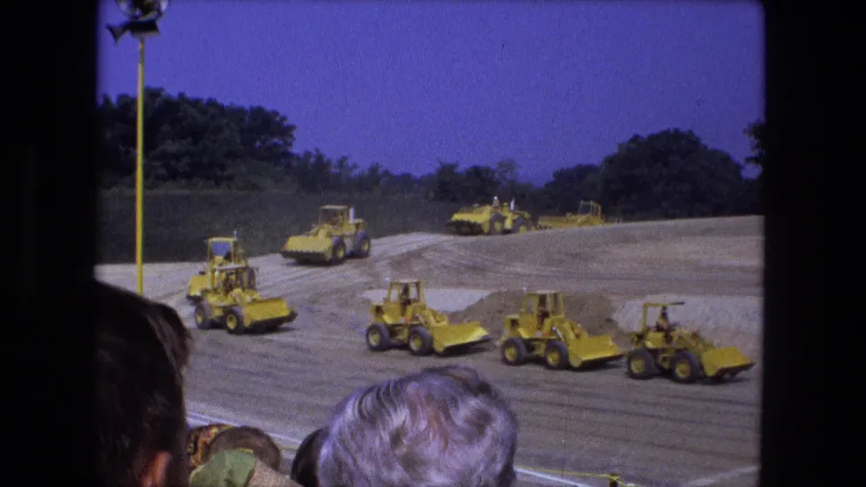 1974 Fort Wayne 8mm frame of yellow earthmoving vehicles crossing a dirt construction site while spectators watch nearby.