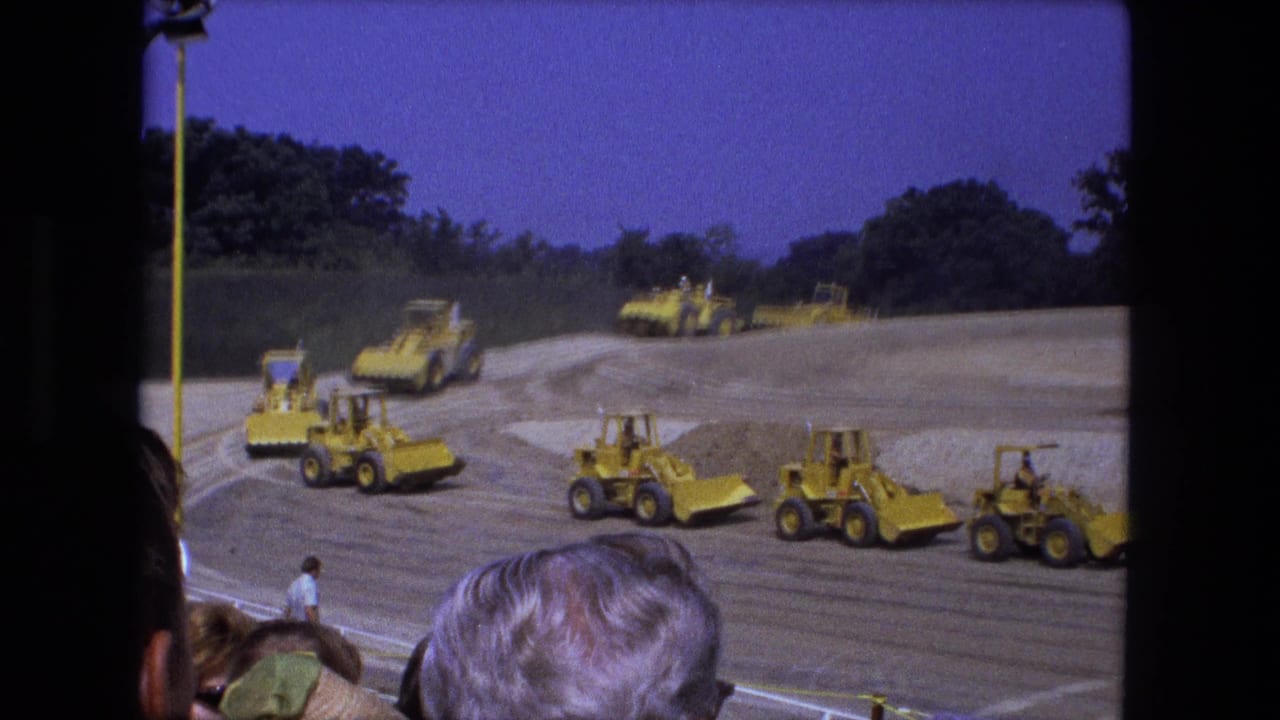 Several yellow construction machines move across a graded dirt area as spectators watch from the foreground.