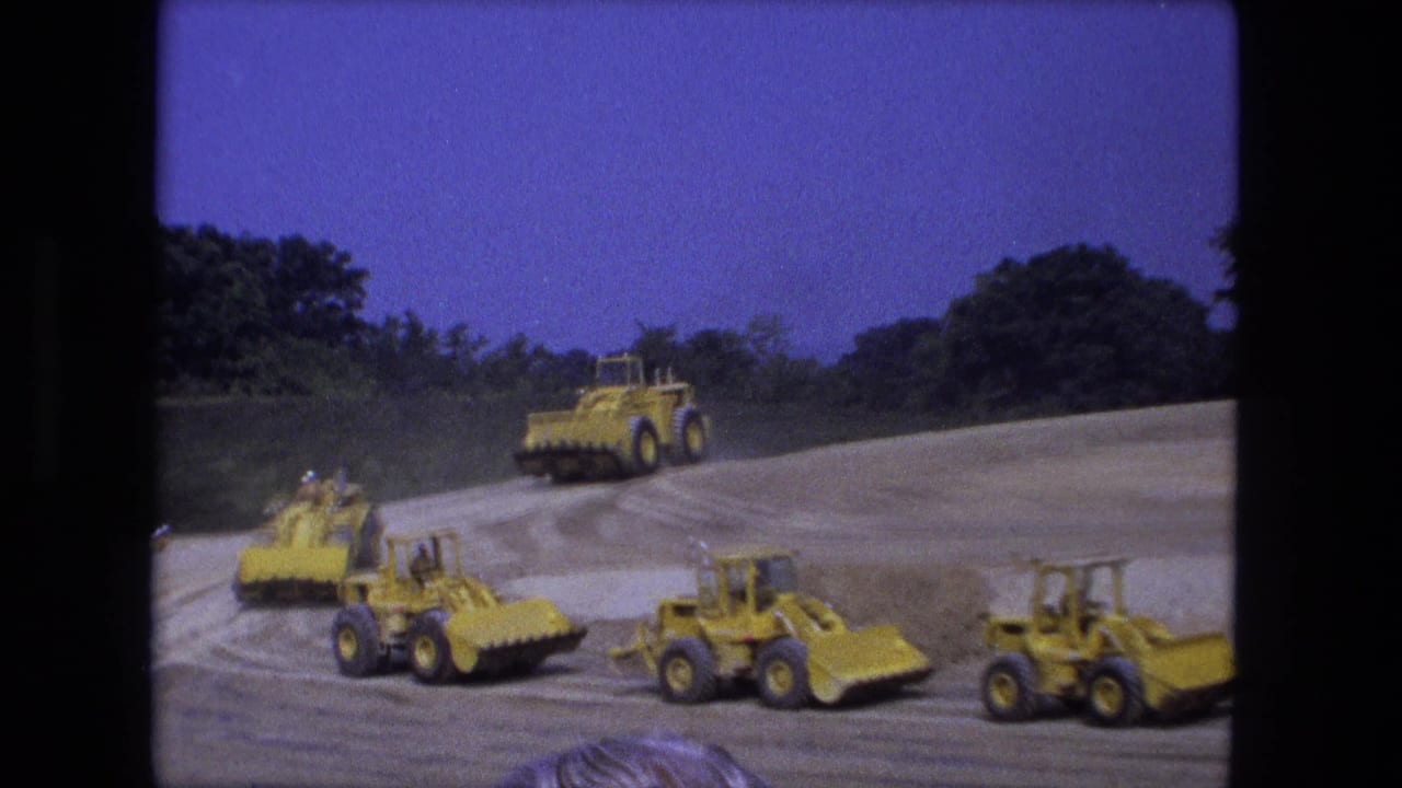 Yellow loaders travel in formation across a dirt worksite bordered by trees under a blue sky.