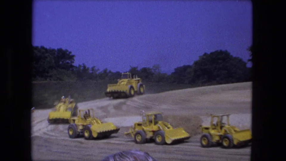 1974 Fort Wayne home-movie view of yellow earthmoving equipment driving across a graded dirt area with trees in the background.