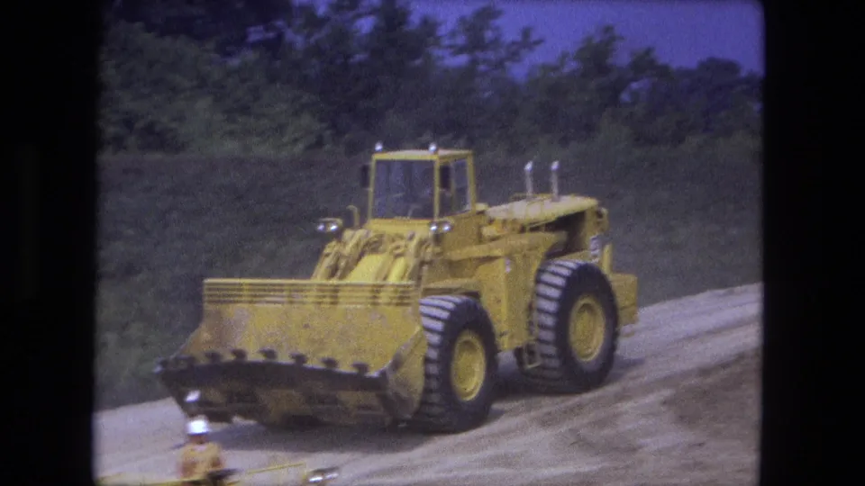 Close Fort Wayne 1974 home-movie view of a large yellow loader traveling on a curved graded dirt roadway with trees behind it.