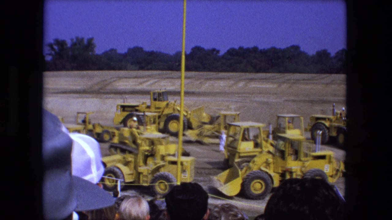 Yellow construction machines operate on a dirt field as spectators watch from behind a barrier.