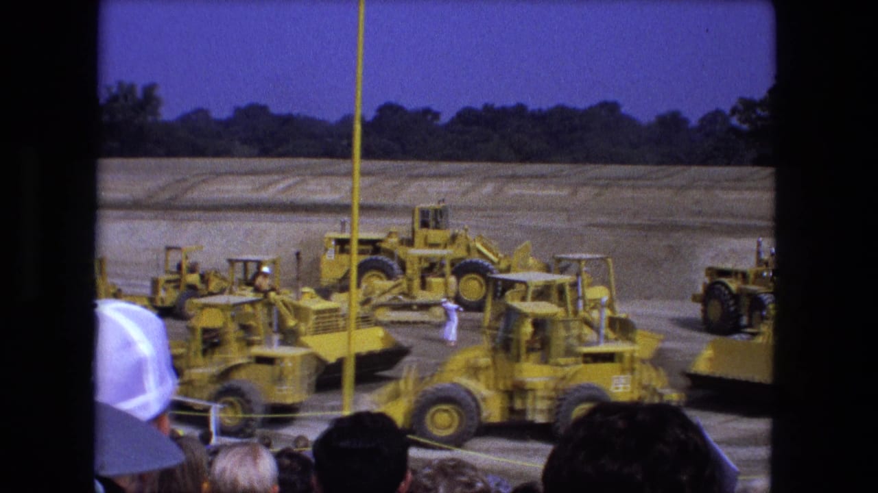 1974 Fort Wayne frame with multiple yellow construction vehicles, workers, a rope barrier, and heads of spectators at the bottom edge.