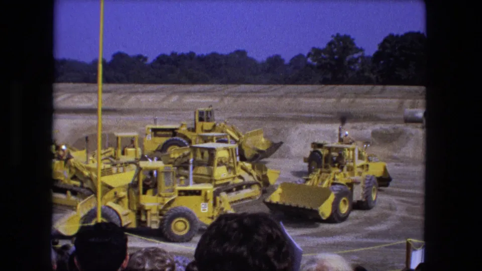 1974 Fort Wayne 8mm frame showing yellow construction equipment passing a roped viewing area with spectators partly blocking the lower frame.
