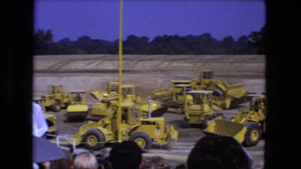 1974 Fort Wayne home-movie frame of yellow loaders and bulldozers arranged on a dirt field during a public equipment demonstration.