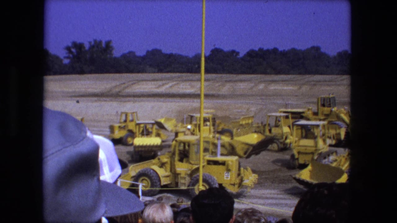 Spectators watch several yellow loaders moving across a dirt construction area.