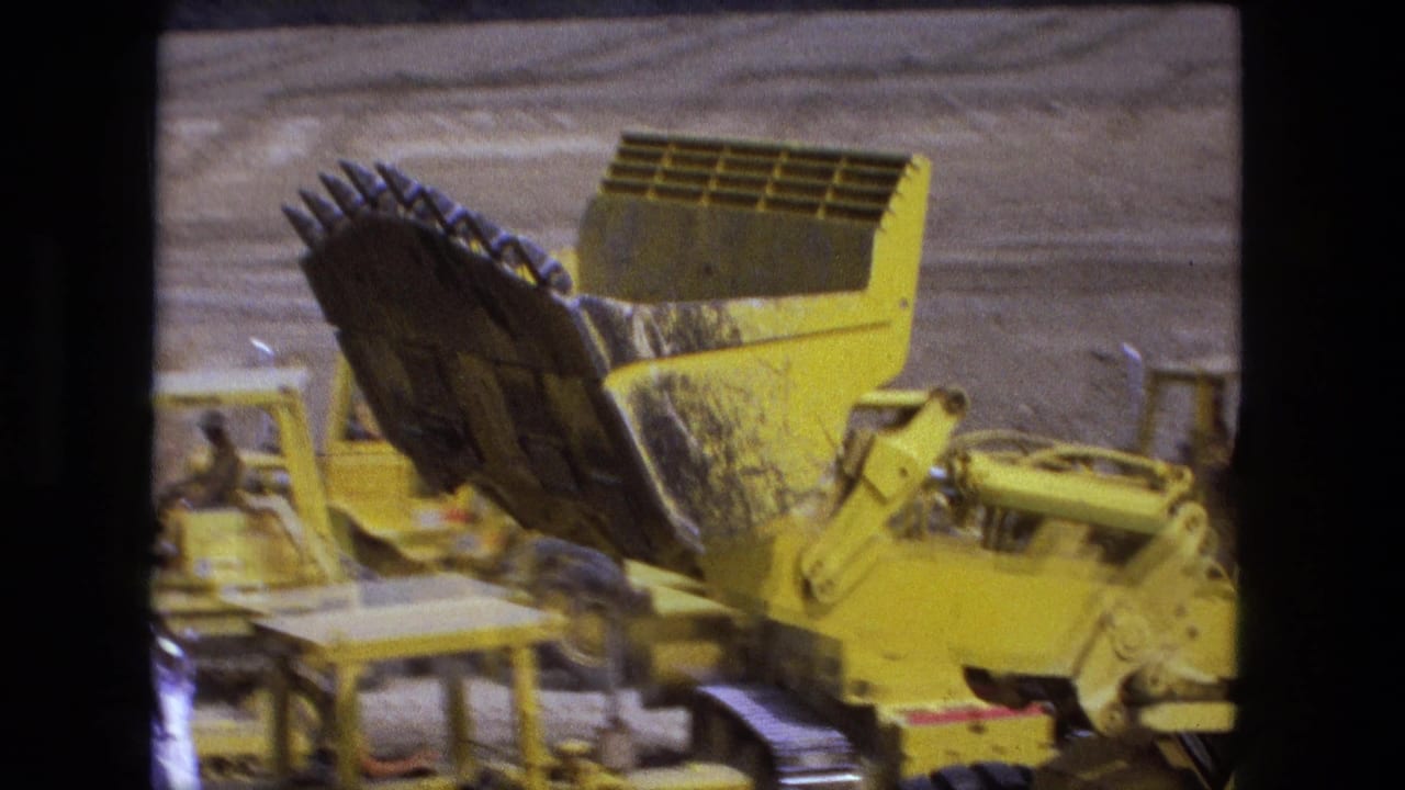 A yellow front-end loader with its bucket raised moves across the Fort Wayne dirt site in 1974 footage.
