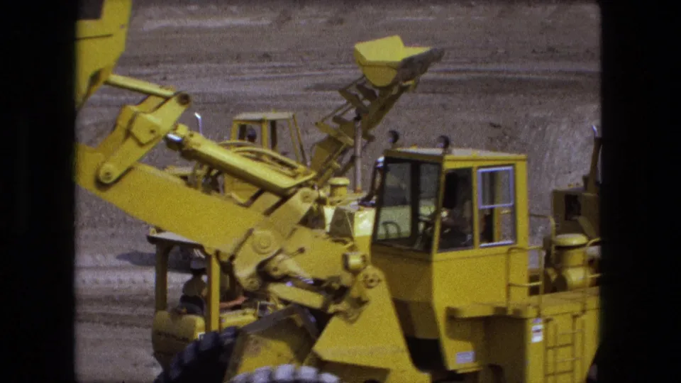 A 1974 Fort Wayne home-movie frame shows a yellow loader arm and bucket raised beside another cab on a dirt lot.