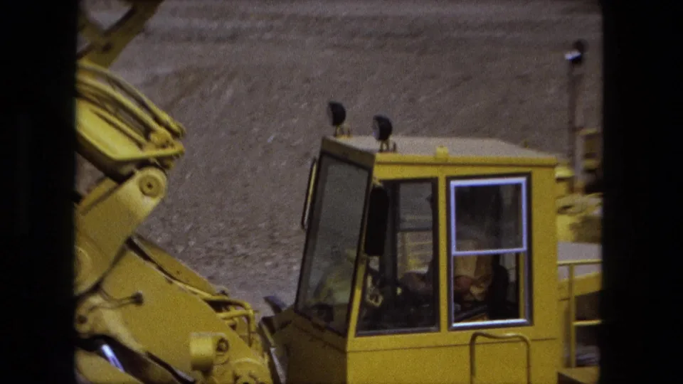 Close Fort Wayne 1974 frame of a yellow loader cab, hydraulic arm, and dirt embankment in handheld 8mm footage.