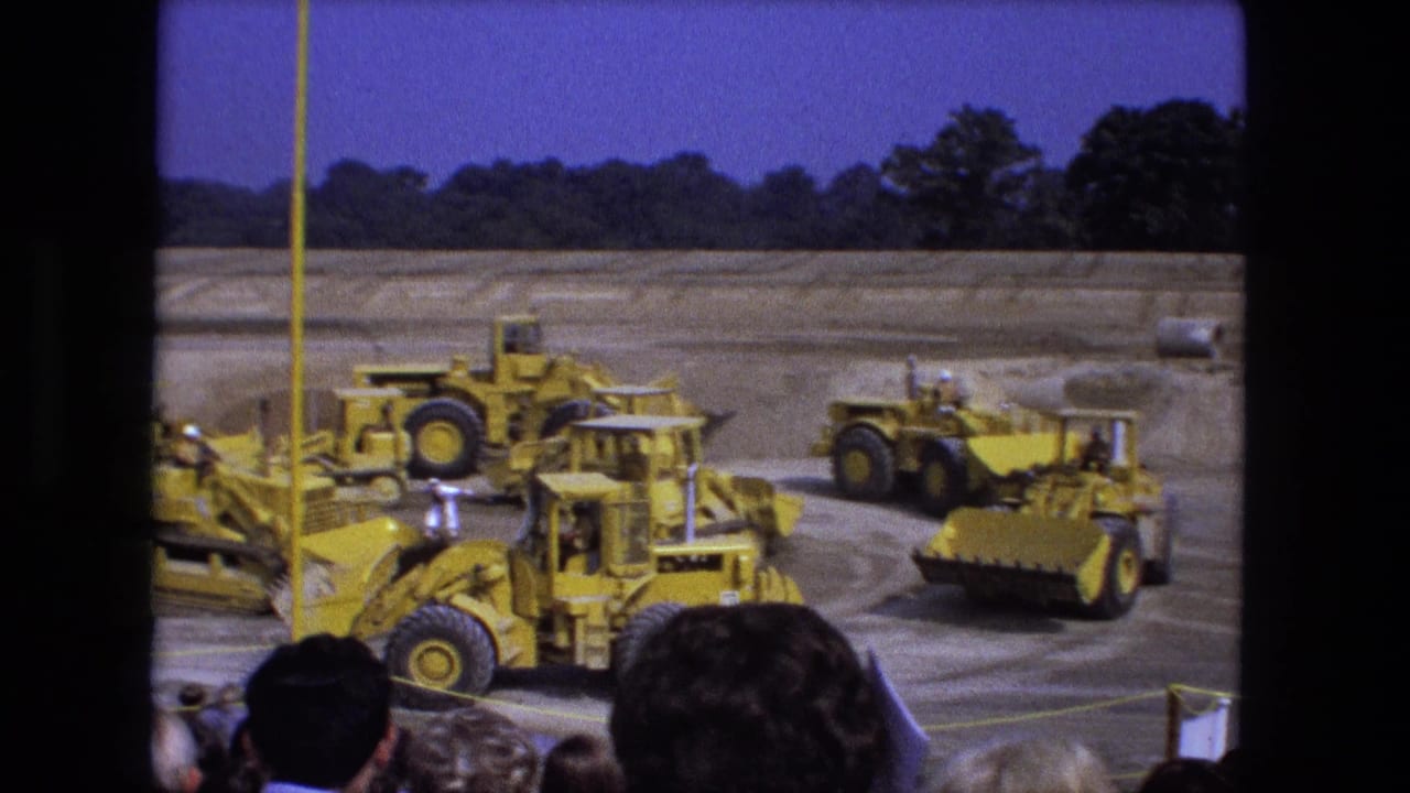 Yellow construction vehicles line up and move on a dirt field behind spectators.