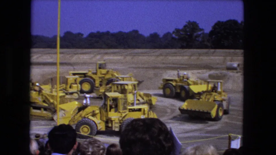 1974 Fort Wayne 8mm view of yellow tractors and construction vehicles on a dusty field with spectators in the foreground.