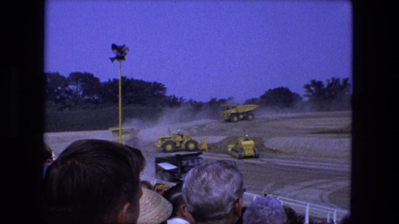1974 Fort Wayne 8mm frame showing a yellow dump truck and smaller machines moving through dust before a crowd.