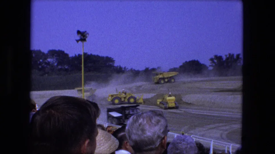 1974 Fort Wayne 8mm frame showing a yellow dump truck and smaller machines moving through dust before a crowd.