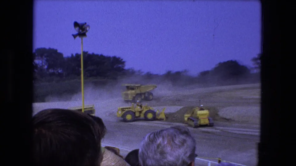 Crowd-level 1974 Fort Wayne 8mm view of yellow heavy equipment on a dusty track with a floodlight overhead.