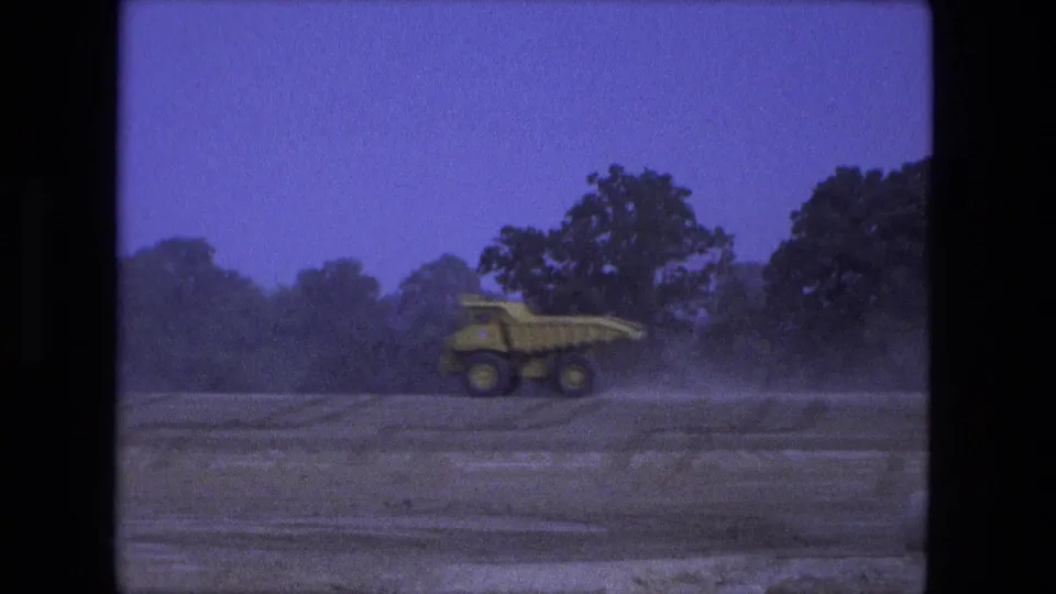 1974 Fort Wayne 8mm frame of a yellow dump truck moving alone across a dusty dirt field under blue sky.