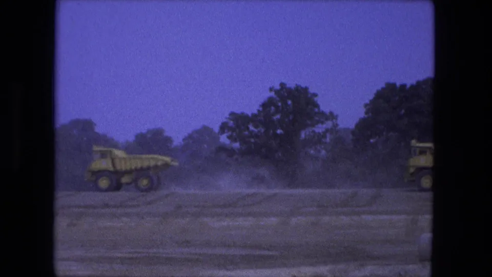 1974 Fort Wayne 8mm frame showing a yellow dump truck moving across dusty ground with a tree line under a purple-blue sky.