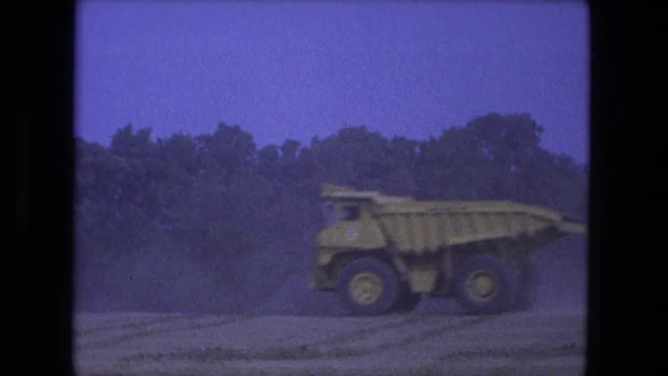 1974 Fort Wayne 8mm frame showing a yellow dump truck moving left across a dusty construction road, softened by motion and grain.