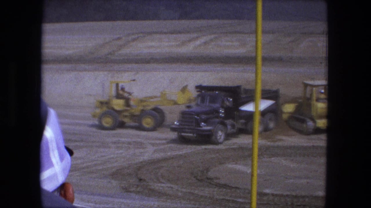 A yellow front loader works near a black dump truck at a Fort Wayne construction site, with a pole and film borders in the foreground.